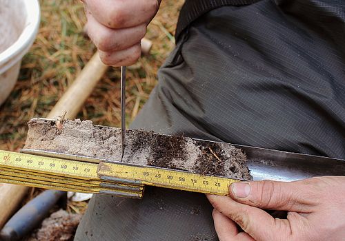 In the center of the picture you can see an auger filled with a soil sample on a person's lap. With one hand, a ruler is placed on the drilling stick, with the other hand the person is cutting off a soil sample with a knife.